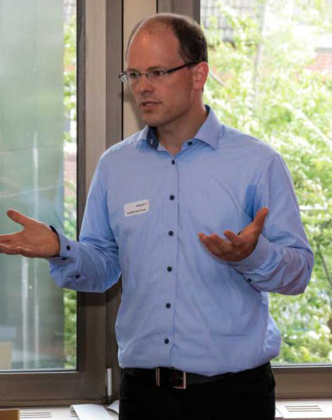 Ein Mann (Trainer Jan-Fredo Willms) mit Brille und Namensschild in blauem Hemd spricht gestikulierend vor einem Fenster, durch das grüne Bäume zu sehen sind.