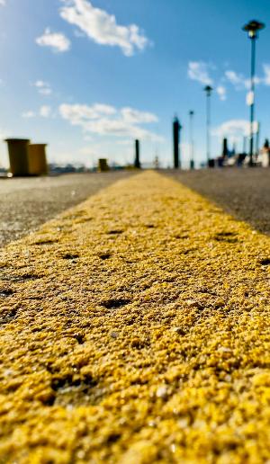Nahaufnahme einer gelben Markierungslinie auf Asphalt an einem sonnigen Tag mit unscharfem Blick auf Hafenanlagen im Hintergrund.