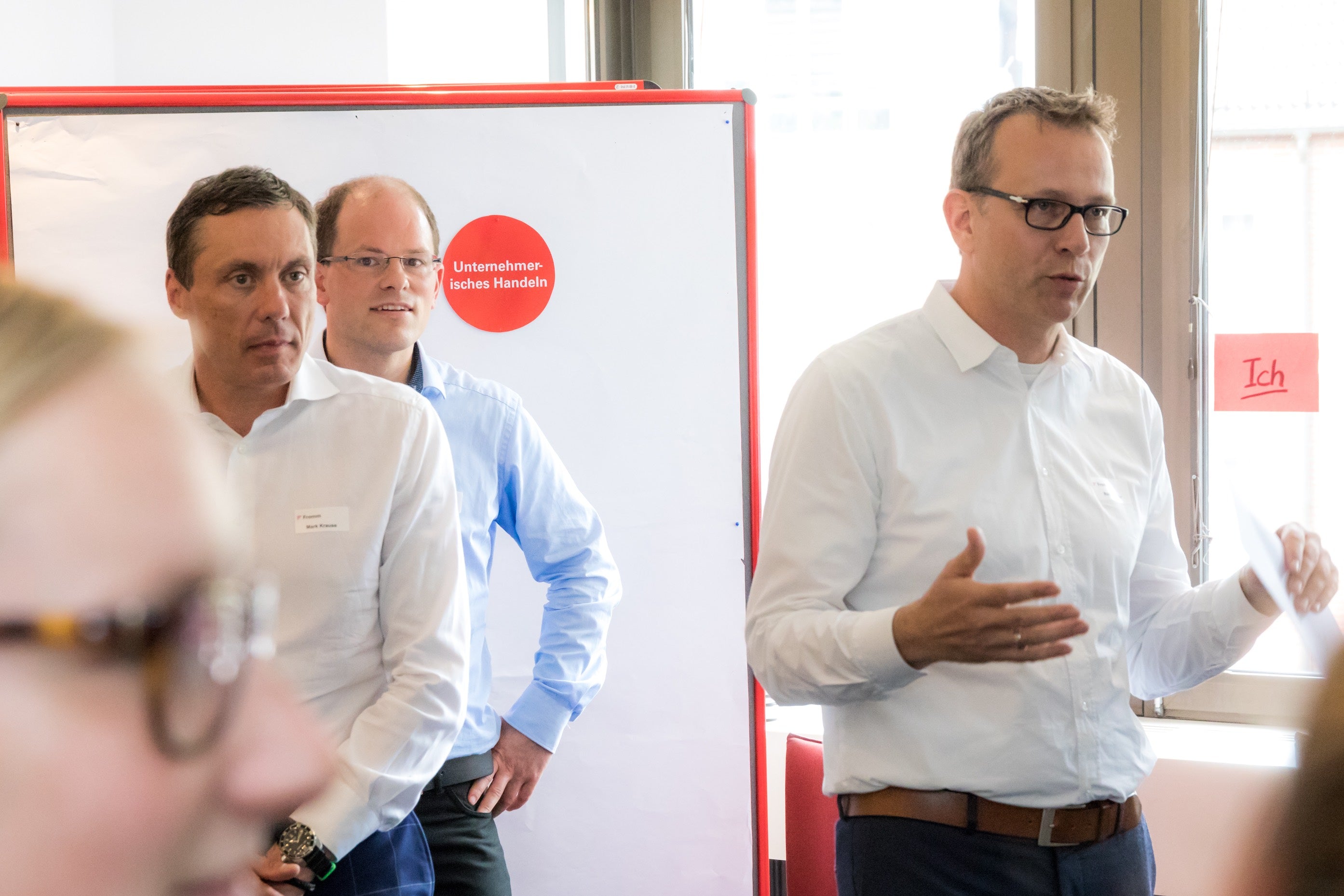 Drei Männer (einer davon Geschäftsführer Ralf Brenner, einer Jan-Fredo Willms) in Hemden stehen in einem Seminarraum vor einem Flipchart mit der Aufschrift „Unternehmerisches Handeln“, während Ralf Brenner spricht und die anderen aufmerksam zuhören.