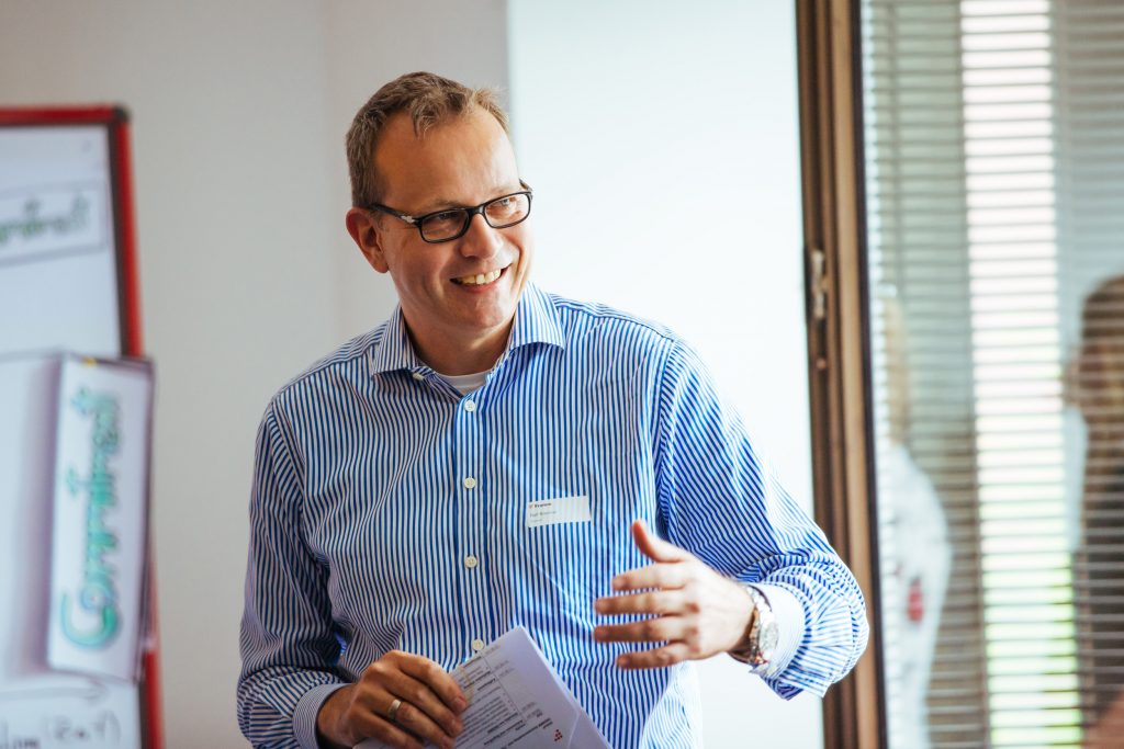 Ein Mann (Geschäftsführer Ralf Brenner) mit Namensschild und gestreiftem Hemd spricht lächelnd vor einem Flipchart und hält dabei Unterlagen in der Hand; im Hintergrund ein Fenster mit Lamellenjalousie.