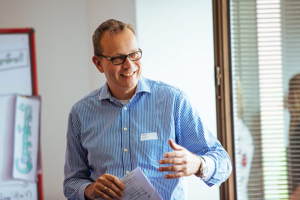 Ein Mann (Geschäftsführer Ralf Brenner) mit Namensschild und gestreiftem Hemd spricht lächelnd vor einem Flipchart und hält dabei Unterlagen in der Hand; im Hintergrund ein Fenster mit Lamellenjalousie.
