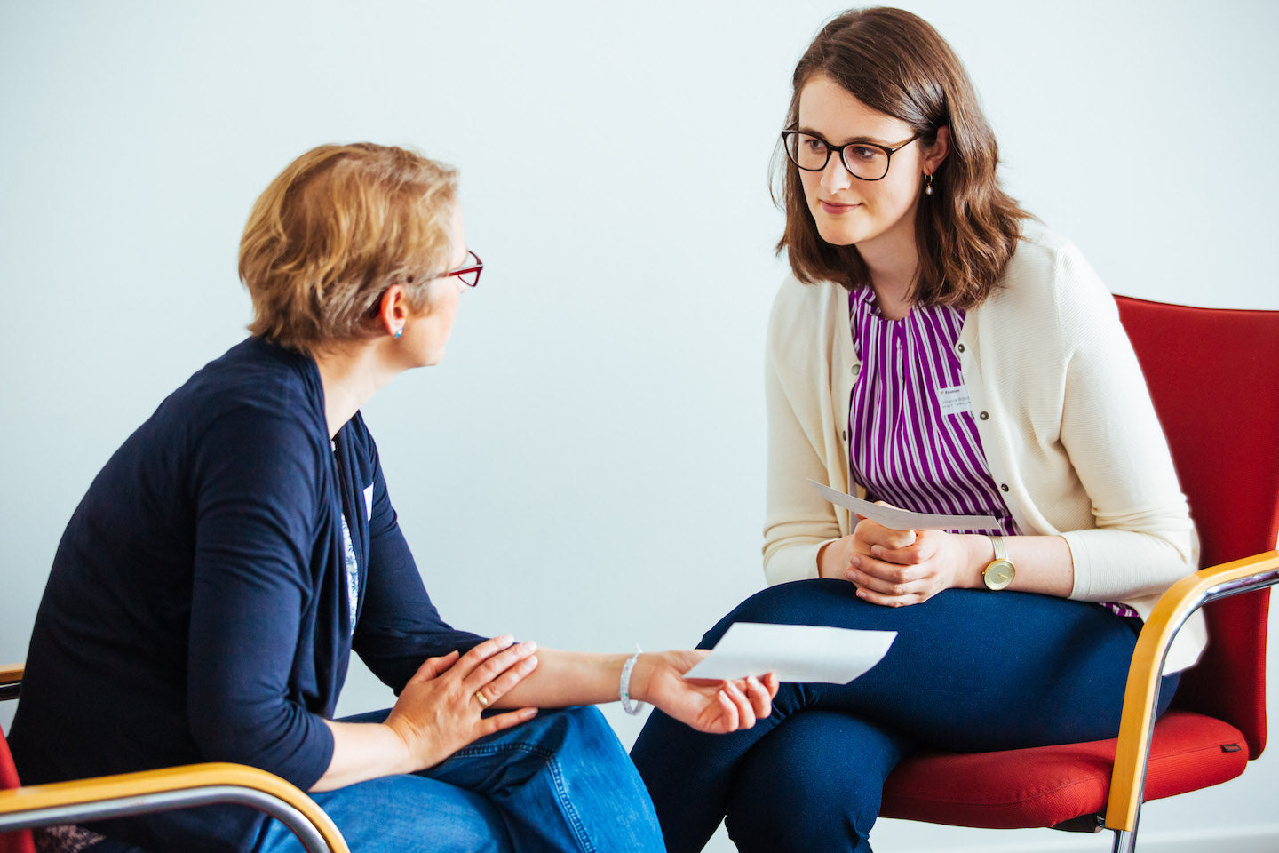 Zwei Frauen (eine davon Trainern Julia Baum) sitzen sich auf roten Stühlen gegenüber und führen ein Gespräch, wobei beide Papierkarten in der Hand halten.