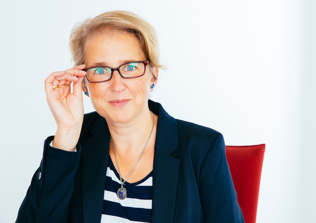 Frau (Trainerin Julia Baum) mit Brille, kurzem blondem Haar und dunkelblauem Blazer sitzt vor weißem Hintergrund auf einem roten Stuhl und blickt direkt in die Kamera.