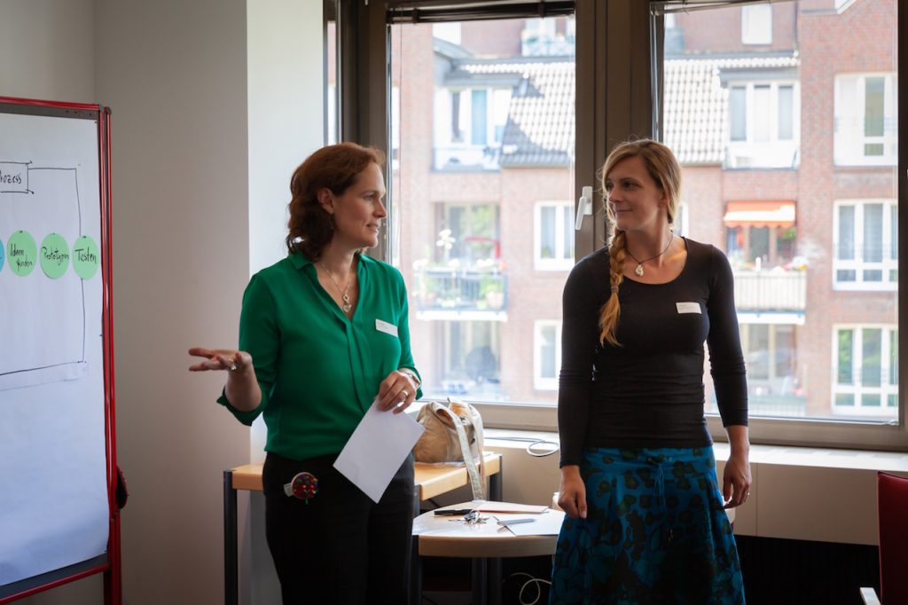 Zwei Frauen (eine davon Geschäftsführung Yvonne Funcke) mit Namensschildern stehen nebeneinander vor einem Flipchart und sprechen, im Hintergrund ein Fenster mit Blick auf Wohnhäuser.