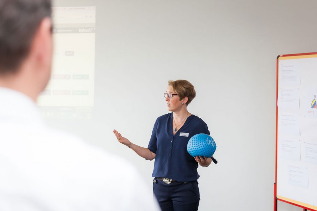 Eine Frau mit Namensschild spricht vor einer Projektionsfläche und hält dabei einen blauen Ball in der Hand, während eine weitere Person im Vordergrund zuhört.