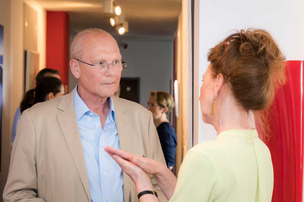 Ein Mann im beigen Sakko und eine Frau (Trainerin Eva-Maria Kerkhoff) im hellgrünen Oberteil stehen einander in einem Flur gegenüber und führen ein Gespräch, im Hintergrund weitere Personen.