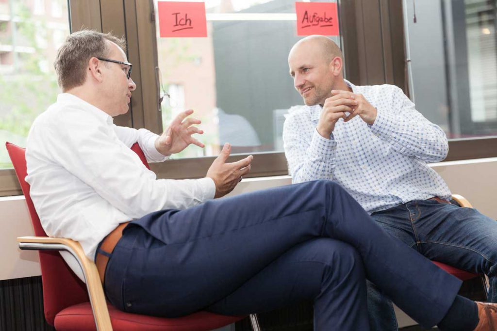 Zwei Männer (einer davon Geschäftsführung Ralf Brenner) sitzen sich in roten Stühlen gegenüber und unterhalten sich intensiv vor einem Fenster, an dem zwei rote Moderationskarten mit den Worten „Ich“ und „Aufgabe“ hängen.