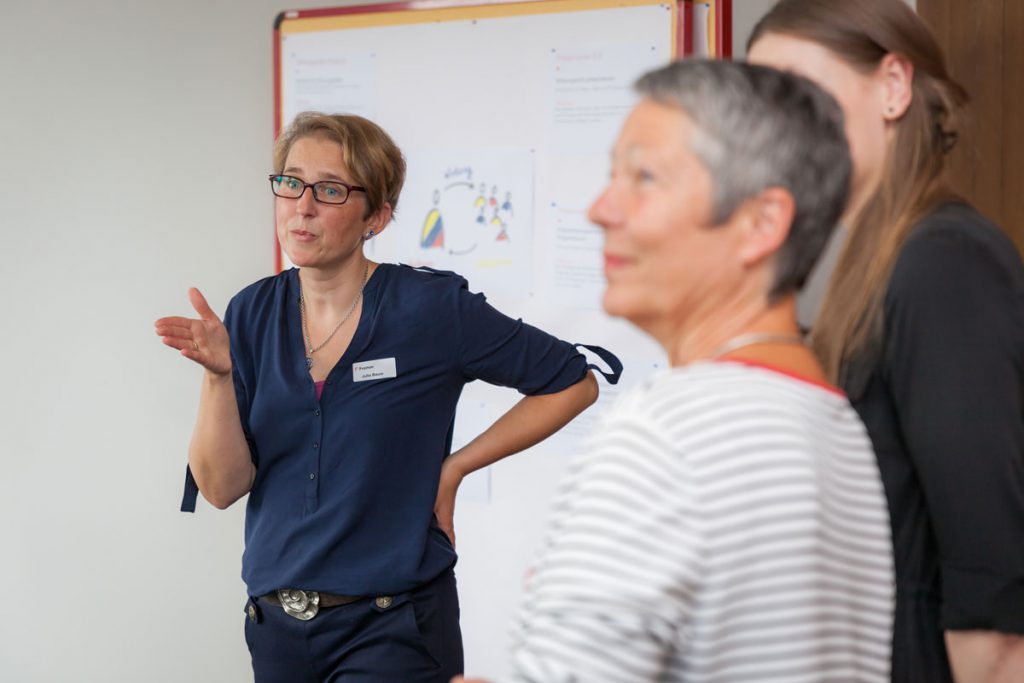 Eine Frau (Trainerin Julia Baum) mit Namensschild spricht vor einer Pinnwand mit mehreren Informationsblättern, während zwei weitere Teilnehmerinnen im Vordergrund zuhören.