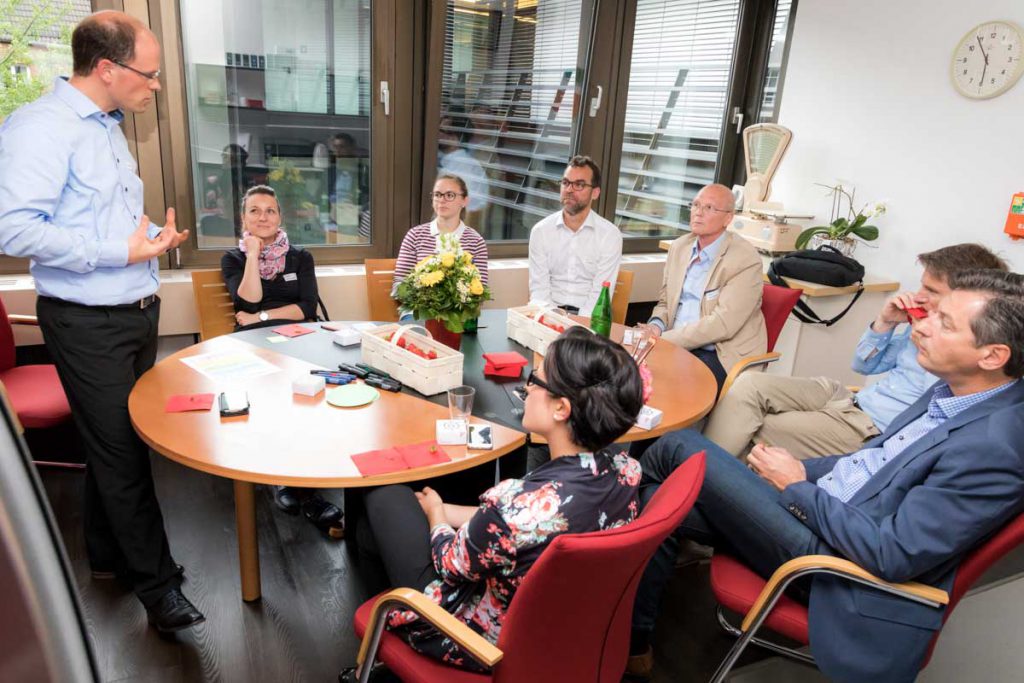 Ein Mann steht vor einem runden Besprechungstisch und spricht zu sieben sitzenden Personen, die aufmerksam zuhören; auf dem Tisch liegen Moderationsmaterialien und es steheen Getränke, ein Erdbeerkorb und ein Blumenstrauß darauf.
