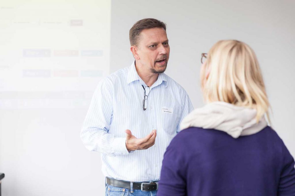 Ein Mann mit Namensschild spricht gestikulierend mit einer Frau, im Hintergrund ist eine Wand mit einer projizierten Präsentationsfolie zu sehen.