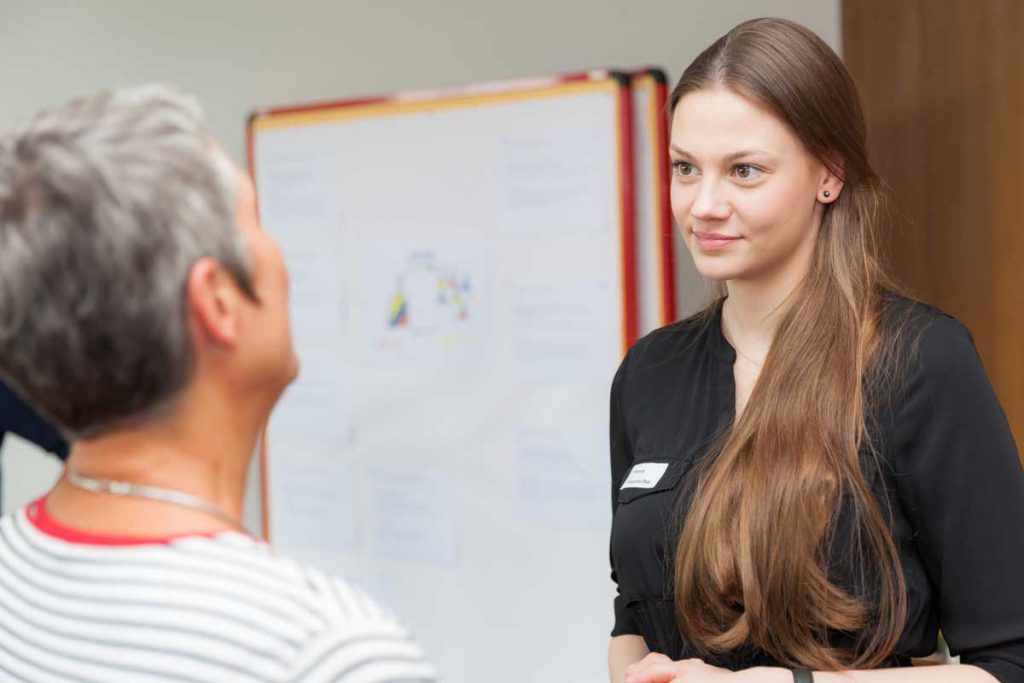 Zwei Frauen stehen sich im Gespräch gegenüber; die eine trägt ein Namensschild und ein schwarzes Oberteil, im Hintergrund ist ein Flipchart mit Texten und Grafiken zu sehen.